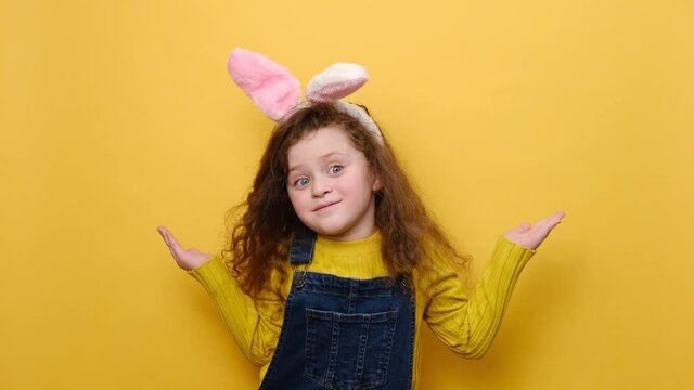 Portrait of confused puzzled preschool little girl kid in pink bunny fluffy ears looking camera spreading hands say oops, isolated over yellow studio background. Easter holiday and emotion concept
