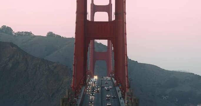 Slow motion traffic on world famous Golden Gate Bridge in pink sunset light. Straight shot of vehicles driving with headlights by the red suspension bridge on in both directions. Modern architecture
