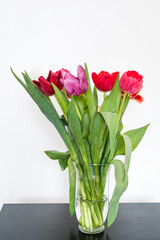 Obraz premium Bouquet of tulips in glass vase on black table. Beautiful spring flowers. White wall. Home decor.