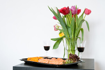 Bouquet of tulips in glass vase. Delicious sushe and rolls set. Japanese cuisine. Couple glasses with wine. Black table. White wall.