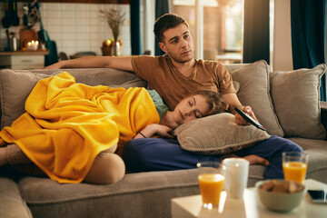 Young man changing channels on TV while his girlfriend is sleeping on the sofa.