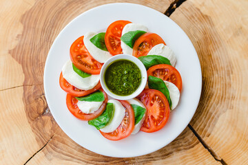Restaurant menu. Sliced tomatoes with mozzarella cheese, basil leaves and pesto sauce in a white plate on a natural wooden background, top view.
