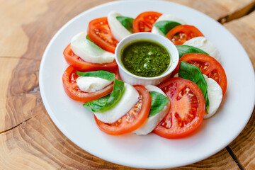 Restaurant menu. Sliced tomatoes with mozzarella cheese, basil leaves and pesto sauce in a white plate on a natural wooden background, top view.