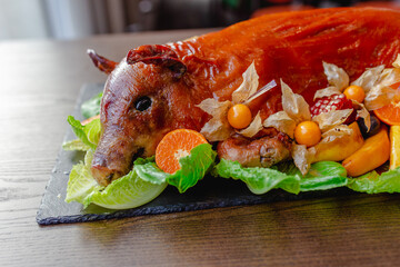 Restaurant menu. Baked suckling pig whole on a stone decorative board with fruit and physalis.  View from the head.