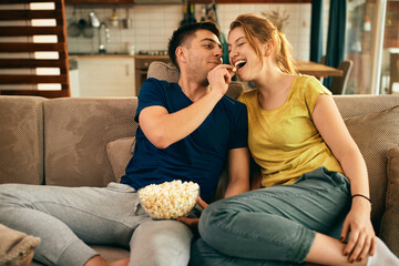 Young man feeding his girlfriend with popcorn while relaxing on the sofa at home.