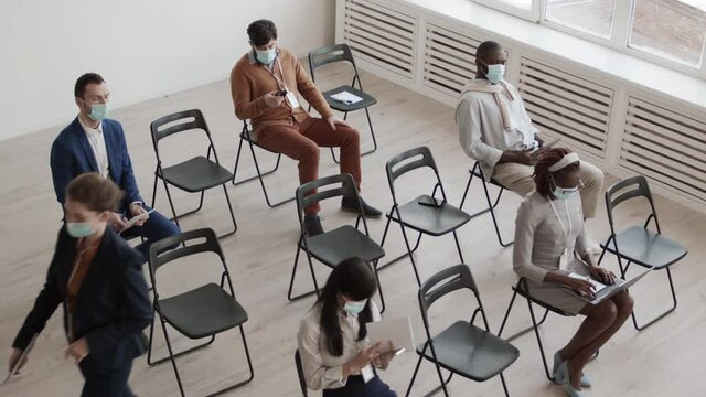 High angle of diverse male and female businesspeople wearing formal clothing and medical face masks, sitting in conference room, keeping social distance, Caucasian woman standing up and leaving
