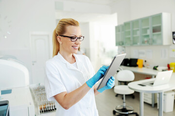 Smiling blond lab assistant with rubber gloves holding tablet and entering test results.