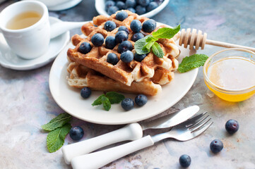 Belgian waffles with blueberries, honey and mint on a white plate. Breakfast with a cup of tea with a sweet waffles. Knife and fork with white handles