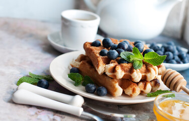 Belgian waffles with blueberries, honey and mint on a white plate. Breakfast with a cup of tea with...