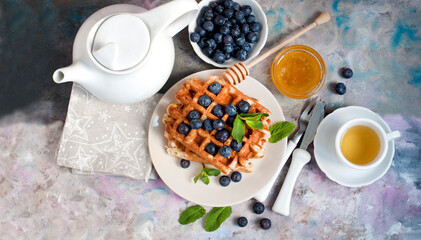 Belgian waffles with blueberries, honey and mint on a white and blue plates. Breakfast with a cup of tea with a white teapot and sweet waffles. Knife and fork with white handles. Top view