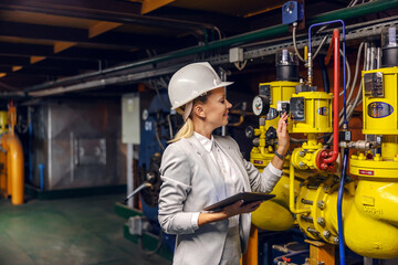 Fototapeta premium Blond dedicated successful businesswoman in formal wear checking on machinery and holding tablet while standing in power plant.