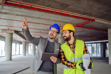 Construction worker and architect standing in building in construction process and discussing about project. Architect pointing at something.
