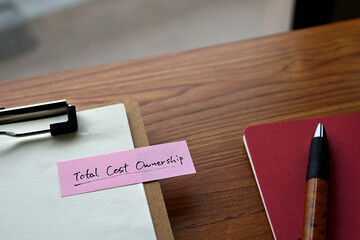 There's a notebook,and a clipboard with a sticky note stuck to it that says Total Cost of Ownership.