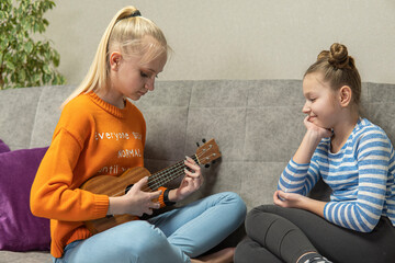 two teenage girls sit on the couch and play the Hawaiian ukulele.