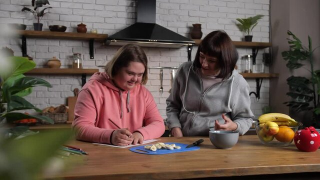 Positive handicapped teenage girl drawing with pencils at kitchen table while curious mother trying to spy. Joyful teenager with down syndrome having fun while hiding drawing process from own mom