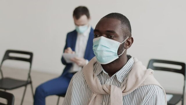 High angle medium close-up of young African male office worker wearing medical face mask, sitting in conference room, looking forward, nodding