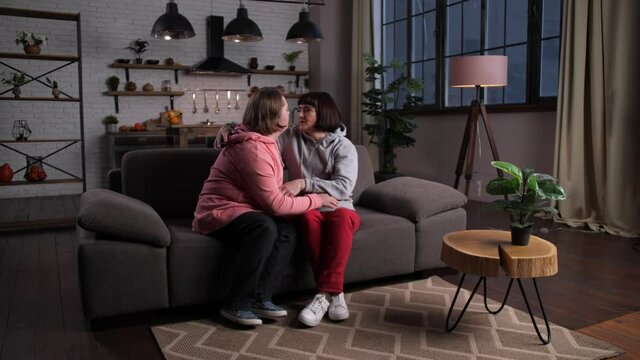 Loving mom leaving tablet and cuddling with disabled teenage daughter while sitting on sofa at home. Happy mother and child with down syndrome expressing love while kissing and embracing each other