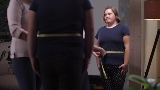 Losing weight disabled teen girl with down syndrome rejoicing at decrease in body weight during healthy diet. Joyful overweight teenager standing by mirror while mom taking measurements of her waist