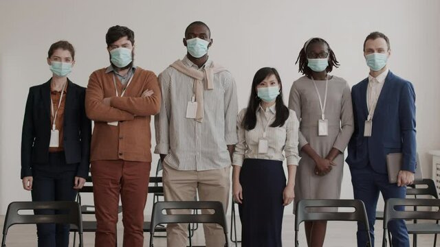 Medium long POV of Asian, African, Caucasian business men and women wearing medical face masks, standing in row in conference room, looking on camera, posing