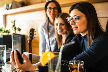 Side view of adult caucasian woman with eyeglasses holding mobile phone while sitting at cafe or restaurant with her female millennial friends making a video call on smartphone or taking selfie photo