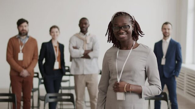 Medium POV of young African female office worker wearing grey dress and badge on neck, looking on camera, standing in foreground of blurred people in conference room