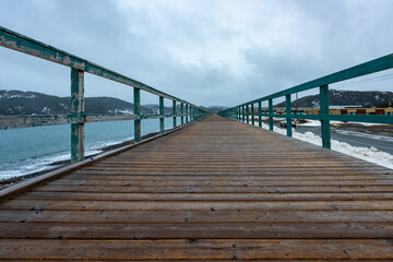A wide worn brown wooden boardwalk with a green wood rail. There are a blue ocean and a treed mountain on one side and snow and a rocky area on the other side.  The sky is covered in grey clouds. 