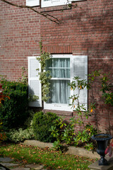 Fototapeta na wymiar The exterior of a red brick wall with a glass porch door, white columns, frame and moulding. There's a single white double hung window with colourful flowers, shrubs and greenery in a flower box. 