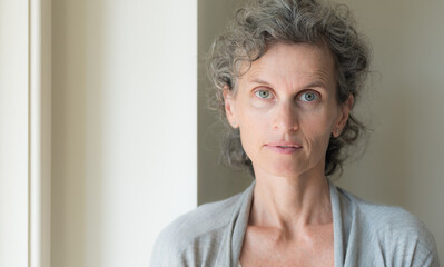 Head and shoulders view of middle aged woman with grey hair looking at camera against beige wall (selective focus)