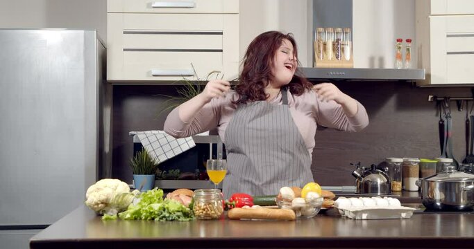 Attractive plus size young woman dancing energetic while cooking in kitchen. Body Positive concept