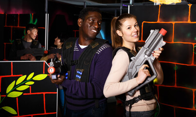 Fototapeta na wymiar happy multiracial couple standing back to back holding laser guns during lasertag game on dark labyrinth