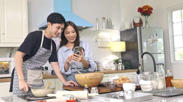 Asian couple Help each other to make a bakery In a romantic atmosphere in the kitchen at home. Young people use smartphones to take pictures while cooking with excited and happy faces.