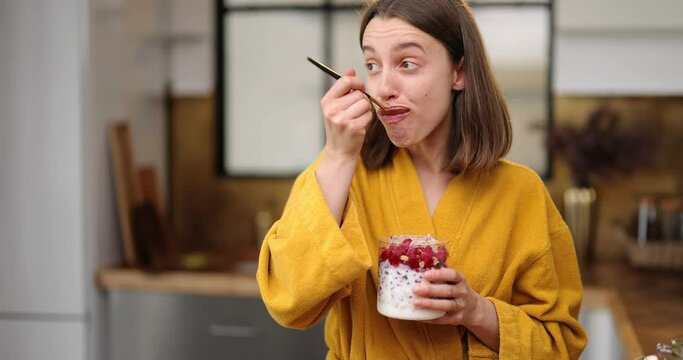 Hungry woman dressed in yellow bathrobe eats yummy cereal breakfast with yogurt and berries from the bowl on the kitchen at home