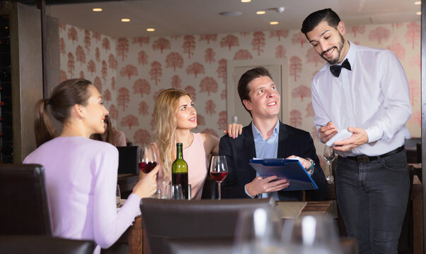Handsom positive smiling waiter taking order from young people in restaurant, writing into note