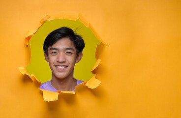 Happy Young Asian teenager man smiling poses through torn yellow paper hole, wearing purple t-shirt with a copy space