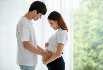 lovely young Asian couple husband and wife standing together smiling and touching a pregnant woman's belly with love and care. Happy and healthy family concept