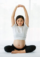 Fototapeta premium healthy mother concept. Young beautiful pregnant woman wearing a white T-shirt and black pants sitting on a bed raises her hands above her head doing stretching after yoga exercise