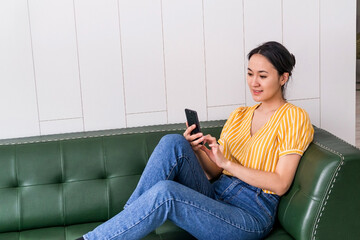 a young attractive Asian woman in a yellow T-shirt is sitting on a green leather sofa with a smartphone in her hands, using the Internet