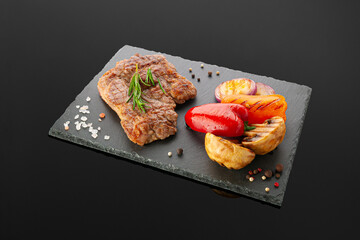 Pork neck steak with grilled vegetables. On a black slate board. Dark background. Isolated.