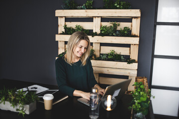 pretty blond young woman is sitting in an ecological office with lots of plants and is working on...