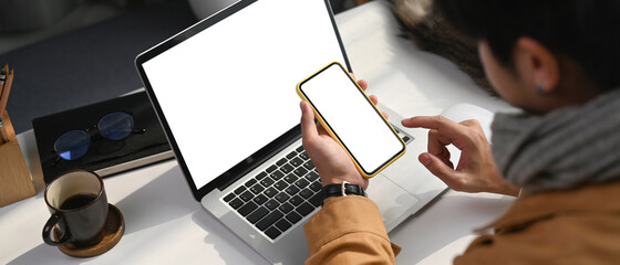 Horizontal photo of young man sitting in front of laptop computer and holding smart phone with...