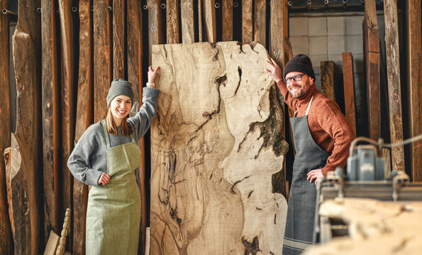 Smiling male and female artisan masters in aprons standing near large lumber plank and looking at camera while working together in carpentry workshop