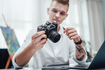 Naklejka premium Professional photographer with tattooed arm looks at photocamera he holding it. Hipster guy wearing formal clothing working in office.