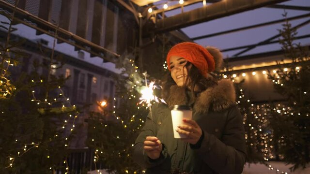 An attractive woman drinks coffee while walking along the street against the background of festive lights of the night city, holding a sparkler in her hands. Glowing blurry bokeh lights of Christmas.