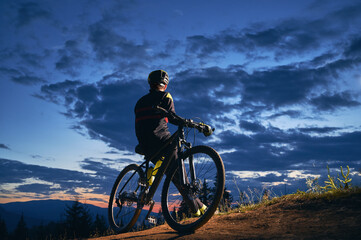 Fototapeta na wymiar Back view of young man in cycling suit sitting on bicycle under beautiful night sky. Male bicyclist in safety helmet resting on hillside road under blue cloudy sky while riding bicycle in the evening.