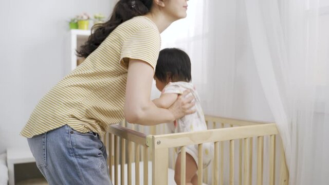 cropped shot asian female parent is returning to pick up her energetic toddler girl and leaving the wooden baby cot in a bright home interior.