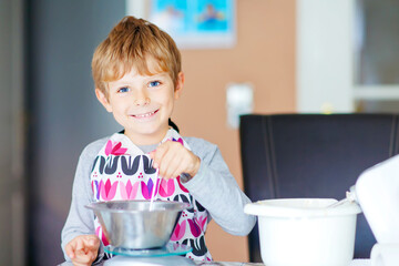 Cute funny blond kid boy baking apple cake in domestic kitchen. Child having fun with helping and working with mixer, flour, eggs and fruits