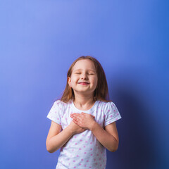 little girl with her eyes closed holds her hands on chest, feels grateful, child dreams with friendly expression, pressing her palms to her chest. concept of love and faith on a purple background