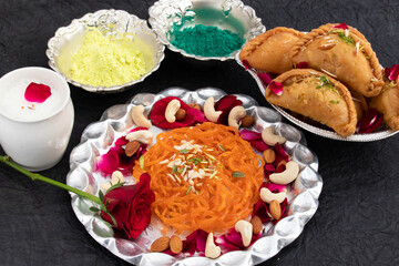Decorative Silver Thali Decorated With Exotic Indian Desserts Jalebi Ghewar Gujia Or Gujiya Mithai Thandai Bhang And Colorful Gulal Abeer Or Abir For Happy Holi On Black Background With Copy Space