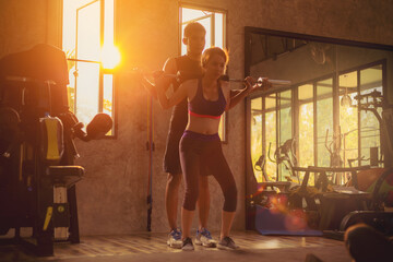 Sporty girl doing weight barbell exercises with assistance of her personal trainer in fitness gym.