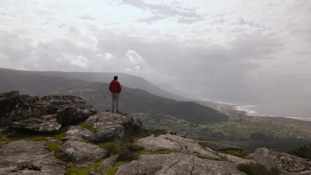 hombre mayor con chaqueta roja y pantalones claros mirando a un horizonte con acantilados, prados, montañas 